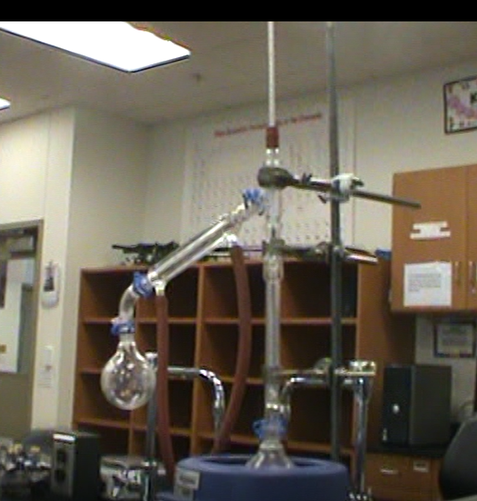 ### Distillation Apparatus in a Laboratory Setting

This image showcases an advanced distillation setup typically found in a chemistry laboratory. This apparatus is used to separate components of a liquid mixture based on differences in boiling points, a fundamental technique in both organic chemistry and industry.

#### Main Components:

1. **Round-Bottom Flask:** The lower left of the image shows a glass round-bottom flask where the liquid mixture to be distilled is initially placed.
   
2. **Heating Mantle:** Located beneath the round-bottom flask, it provides uniform heating to the mixture.

3. **Thermometer Adapter and Thermometer:** This is attached at the top of the apparatus, giving accurate temperature readings to ensure proper monitoring of the boiling point.

4. **Condenser:** Positioned horizontally in the middle and extending downwards, this piece cools the vapor back into a liquid. The cold water enters from the bottom and exits from the top, ensuring efficient condensation.

5. **Clamps and Stands:** These provide structural support for the apparatus, holding the various glass parts securely in place.

6. **Distillation Bridge and Receiving Flask:** The component that connects the condenser and the receiver flask, allowing the condensed liquid to collect in a new container.

7. **Laboratory Setup:** The background includes shelves that likely store additional laboratory equipment and a chart, probably a periodic table, providing quick reference information for chemical properties and elements.

### Educational Importance:

#### Understanding the Distillation Process:
- **Heating:** The substance to be purified is heated gently and gradually. When the temperature reaches the boiling point of the first component, it evaporates.
- **Condensation:** The vapor travels through the condenser, where it is cooled and converted back into a liquid.
- **Collection:** The purified liquid is collected in the receiving flask, while the remaining substance in the round-bottom flask can undergo further distillation if needed.

#### Practical Applications:
- **Purification of Liquids:** Common in both industrial and research environments for purifying chemicals.
- **Separation of Mixtures:** Vital for separating complex mixtures into individual components for further study or use.

Understanding and mastering the use of a distillation apparatus are essential skills for chemistry students and professionals, providing foundational knowledge for many advanced chemical processes and techniques.