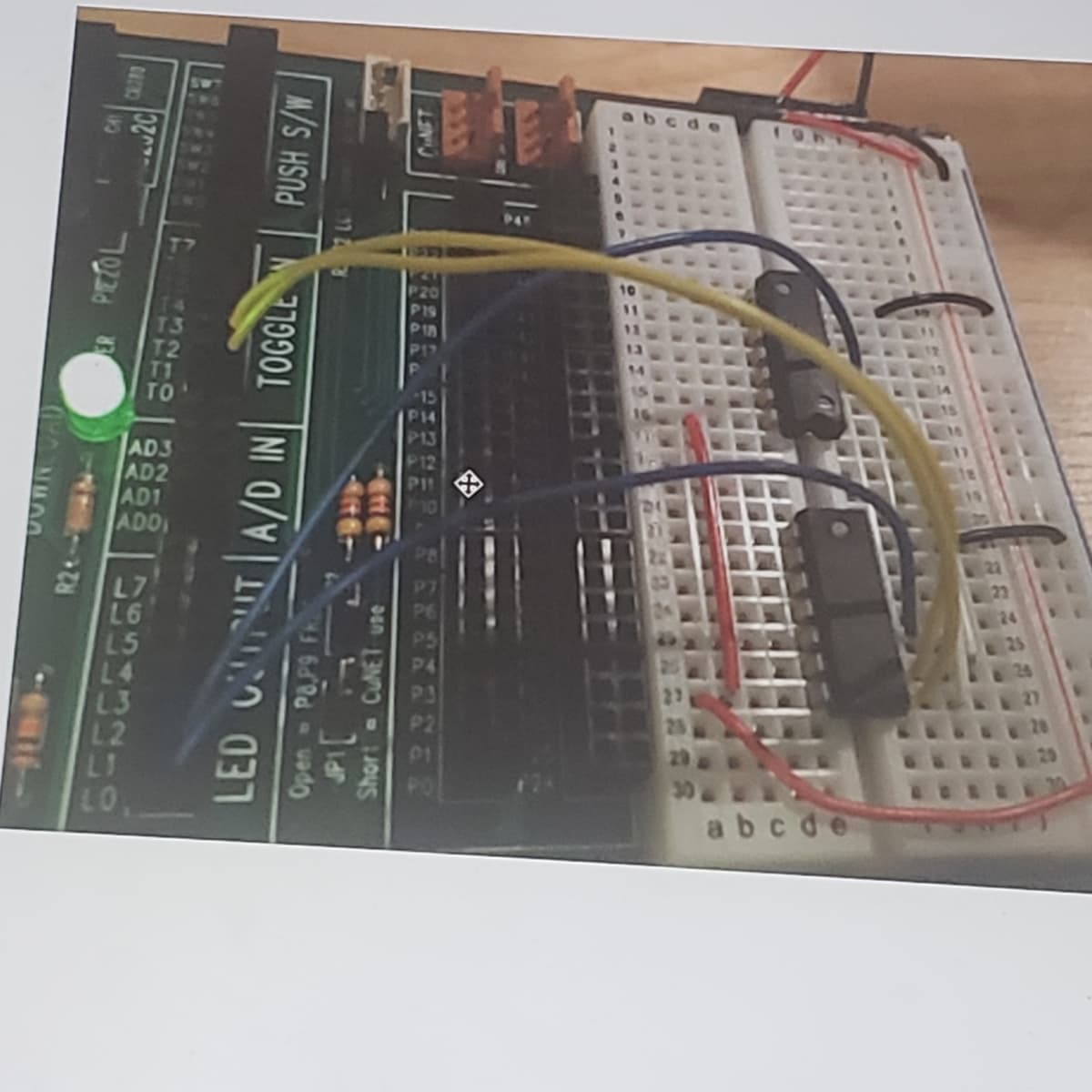 ### Example of LED Interface with Breadboard and Microcontroller

In this educational example, we demonstrate the use of a breadboard connected to a microcontroller development board for controlling LEDs. This setup is ideal for beginners learning about electronics and programming microcontrollers.

#### Components:
1. **Breadboard**: A solderless device used to build electronic circuits. The breadboard used is a typical white breadboard with grid-like holes for component placement.
2. **Microcontroller Development Board**: It has various labeled pins for connecting different inputs and outputs. Visible labels include AD1, AD2, P3, P4, etc. This board is used to interface with the breadboard.

#### Connections:

- **Green LED**: Placed on the development board, it indicates power or specific signal conditions.
- **Resistors**: Provide necessary resistance in the circuit to protect components like LEDs.
- **Wires**: Color-coded wires are used to connect different components.
  - **Yellow Wire**: Connected from the P4 pin on the microcontroller to a component on the breadboard.
  - **Blue Wires**: Used for connections between various points. One connects from below the green LED to the breadboard.
  - **Red Wires**: Power connections, likely connecting the positive rail of the breadboard to the microcontroller.

#### Microchips:
Two integrated circuits (ICs) are mounted on the breadboard, used to manage inputs and outputs or perform specific tasks in the circuit. 

#### Detailed Explanation:
The circuit demonstrates basic interfacing techniques for controlling LEDs using a microcontroller. The steps typically include:

1. **Placing Components**: Place the components such as ICs and LEDs on the breadboard.
2. **Wiring Connections**: Use wires to connect components on the breadboard to the microcontroller board, ensuring correct alignment with the labeled pins.
3. **Power Supply**: Ensure the breadboard is powered correctly, connecting the power rails to the microcontroller’s power source.
4. **Programming**: Upload appropriate code to the microcontroller to control the LED’s behavior based on inputs processed by ICs. 

This setup can be expanded to include more LEDs, sensors, or different input devices, providing a basic yet effective learning platform for understanding circuits and microcontroller programming.